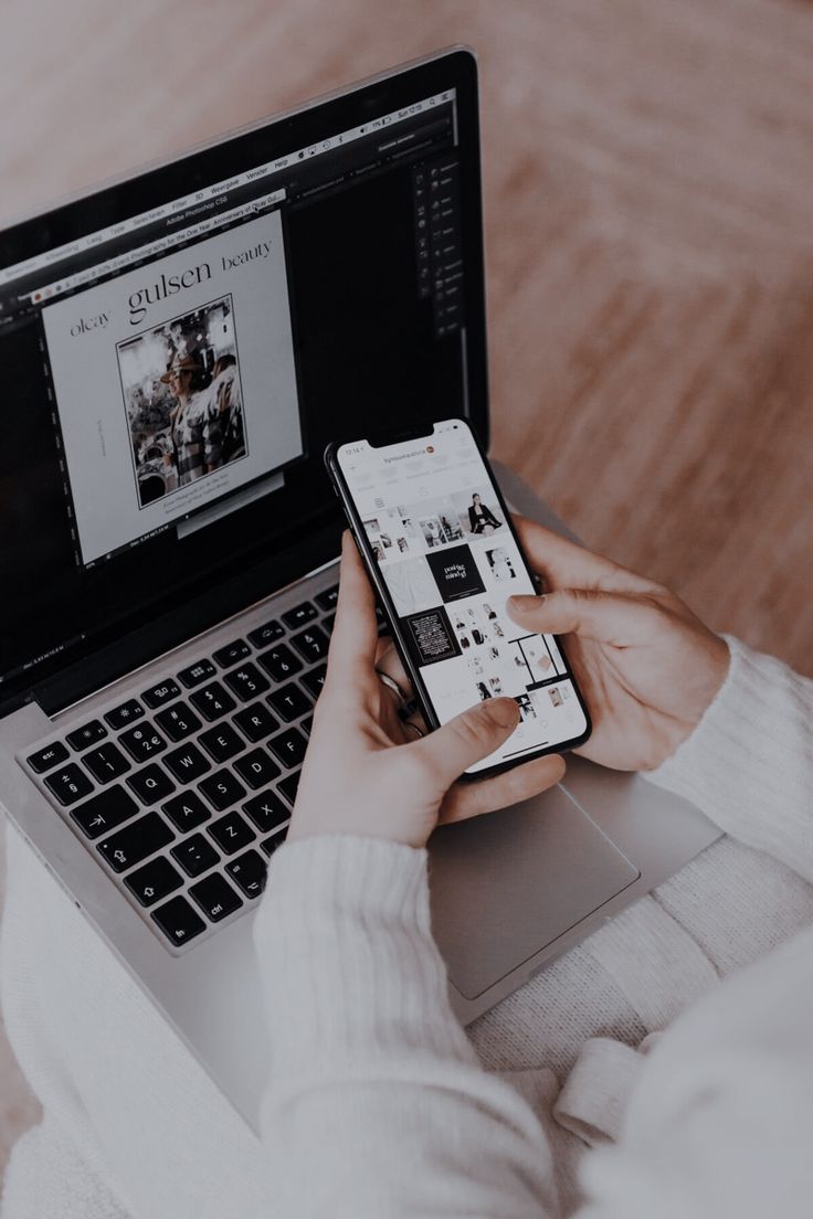 A person in a white sweater using a smartphone while working on a laptop, both displaying digital content and design projects.